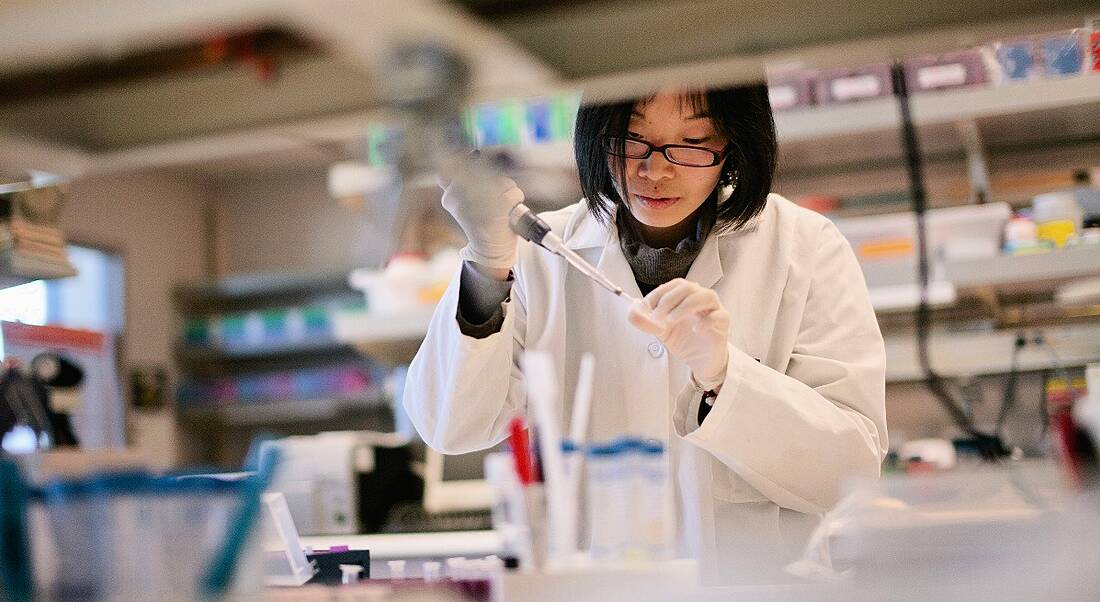 A woman in a white lab coat wearing disposable gloves can be seen pipetting in a lab from across a bench busy with equipment and test tubes.