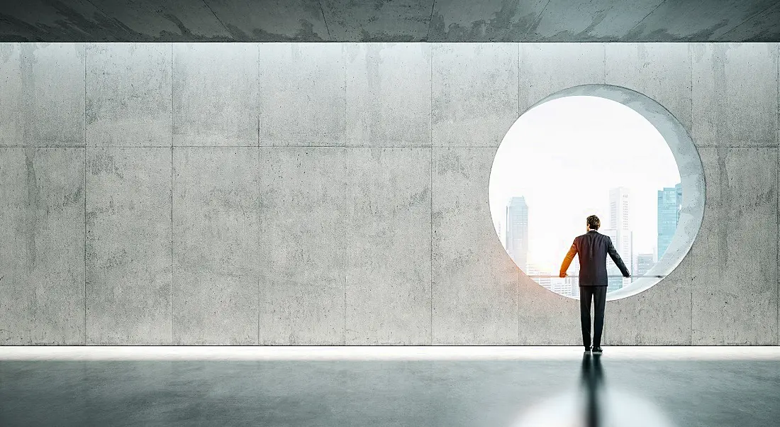 A man stands in a futuristic hallway looking out a large circular window to the city.