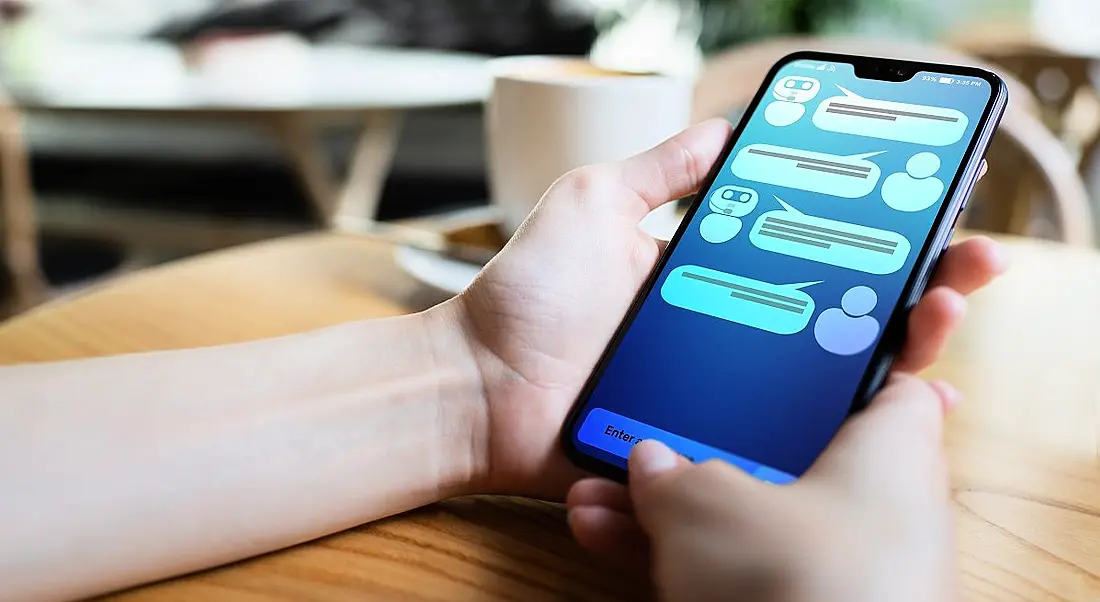 Person holding a mobile phone on a wooden table engaging with a chatbot.