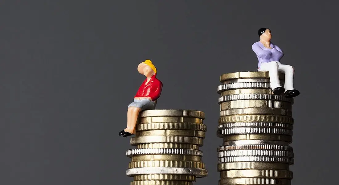 Concept of small figurines of a woman sitting on a smaller pile of coins beside a man on a larger pile of coins.