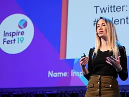 A man in a slate grey suit grinning widely at a crowd at the Inspirefest 2019 conference.