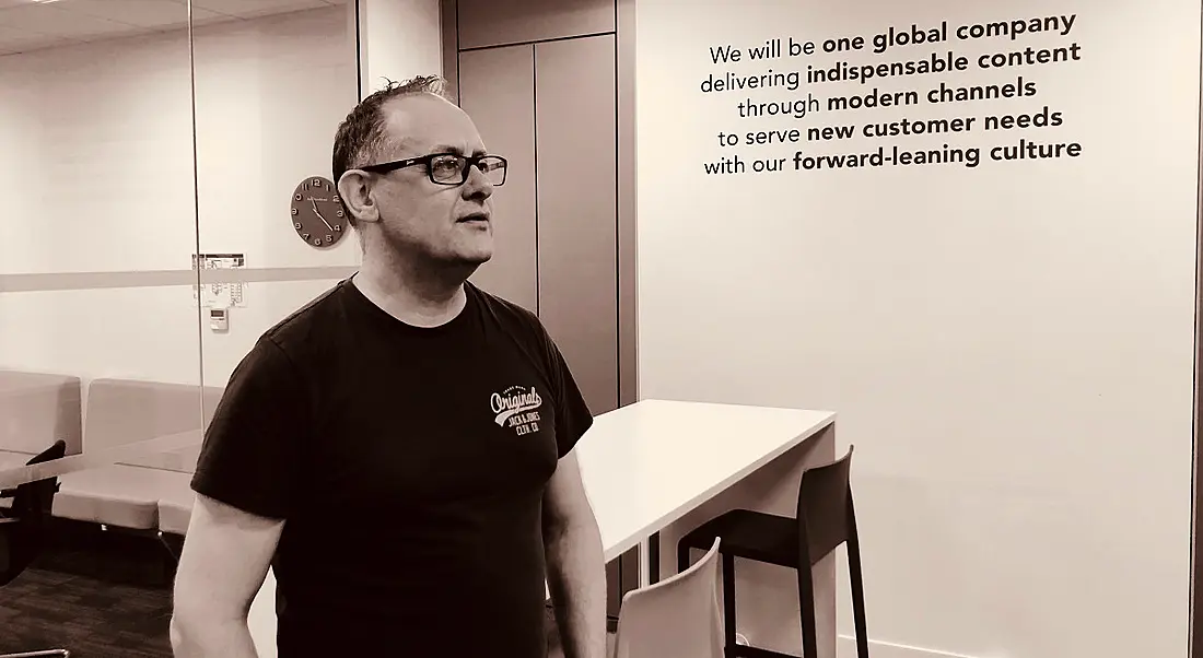 Black and white photo of a man in casual clothes in an office with an inspirational quote on the wall behind him.