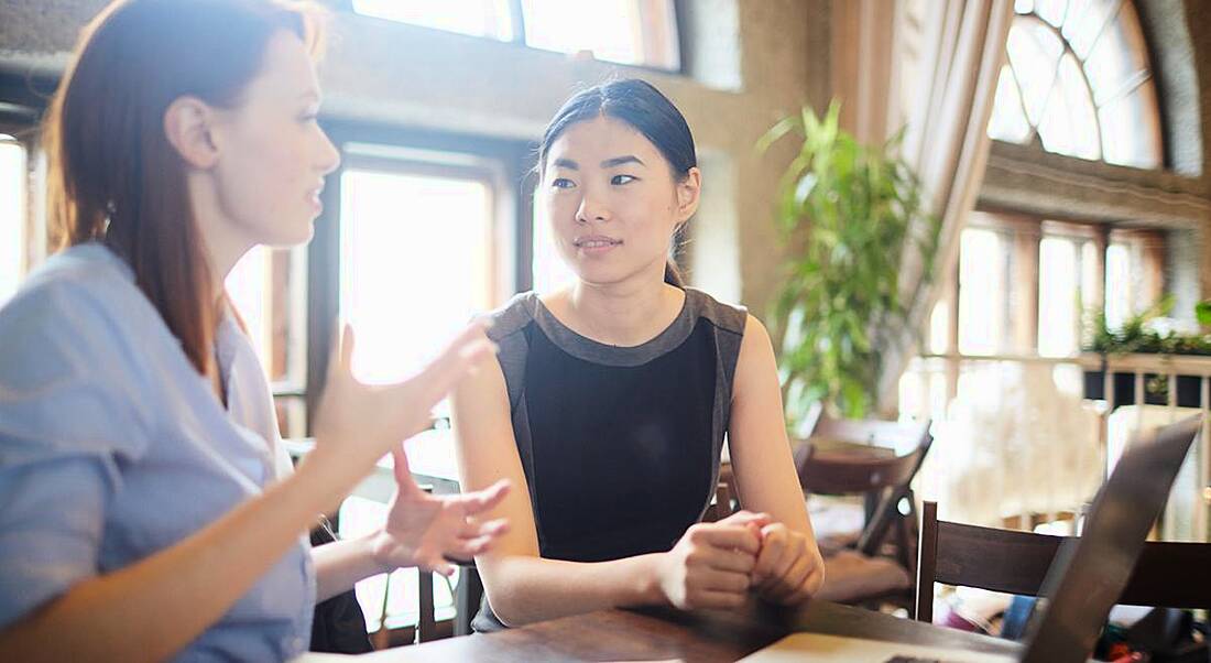 Two women sitting at a table talking in a brightly lit office.