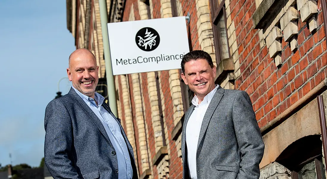 Two men in business attire standing outside Georgian building on sunny day with sign above saying MetaCompliance.