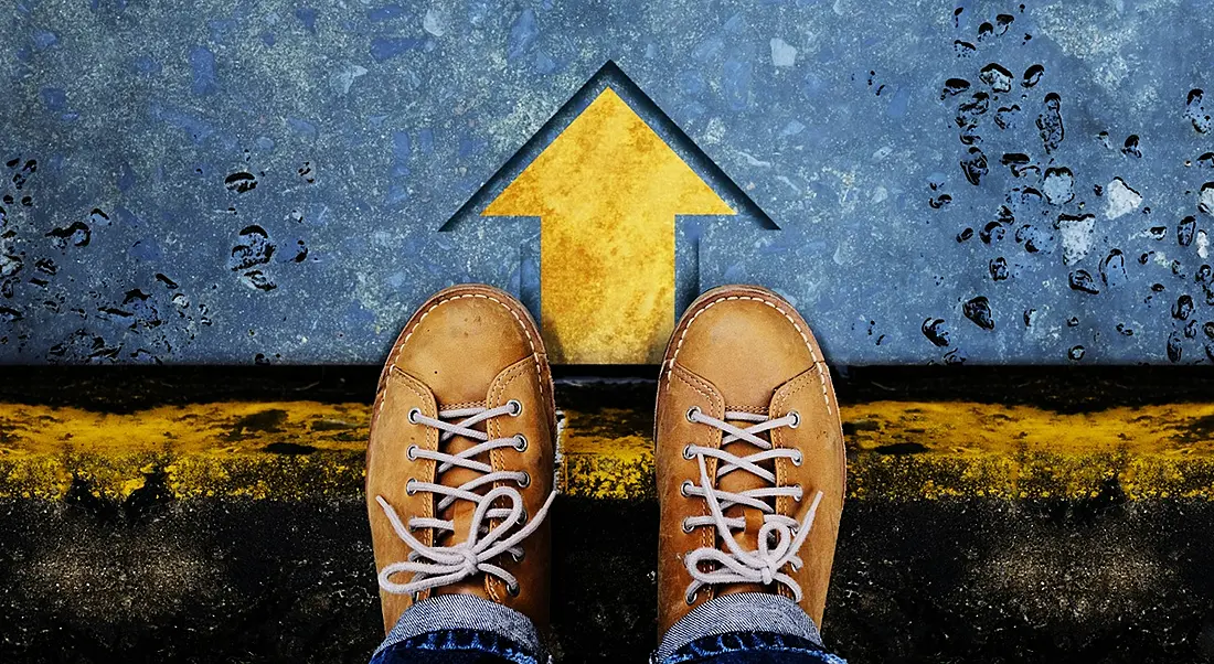 Top view of man with leather shoes standing on a crossroad with an arrow pointing forward.