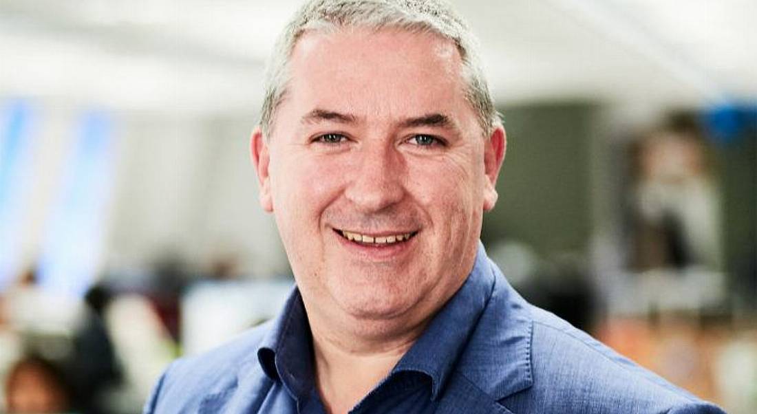 View of man with grey hair smiling at camera wearing dark blue shirt with matching blazer. Blurred background of office.