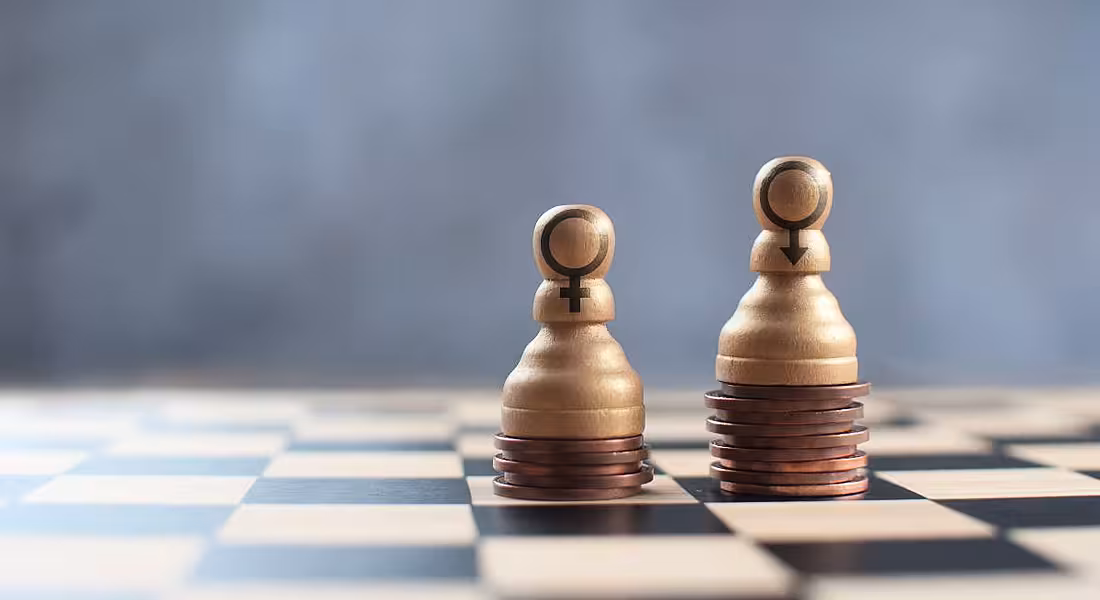 Two chess pawns on a chess board, stacked on top of a heap of coins with varying heights.
