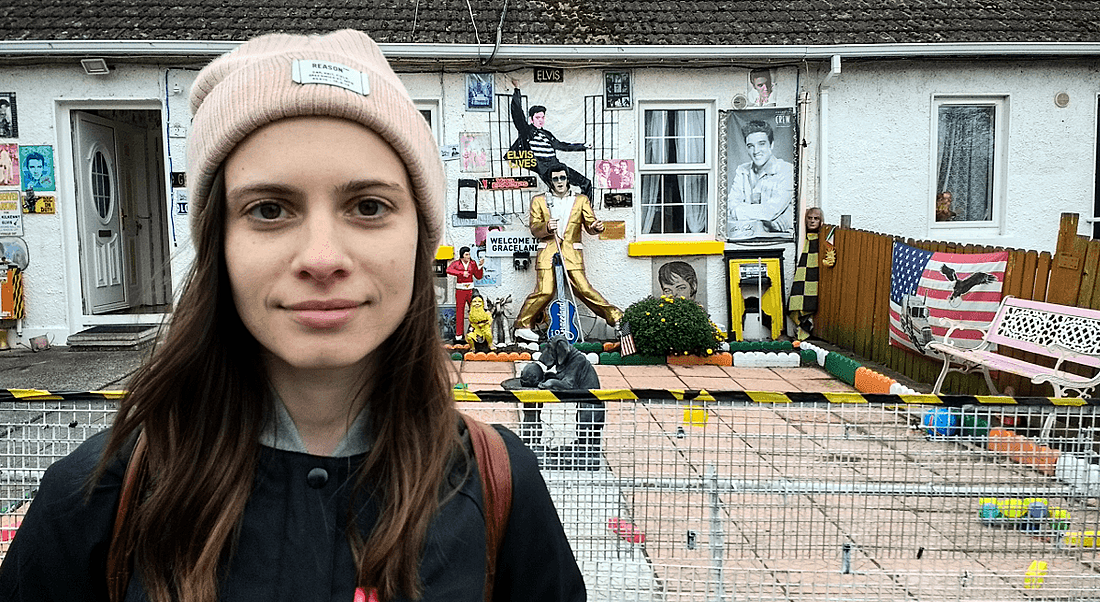 View of young woman in beanie hat standing in back patio of garden on overcast day. Behind her the house and garden are decorated with Elvis paraphernalia.