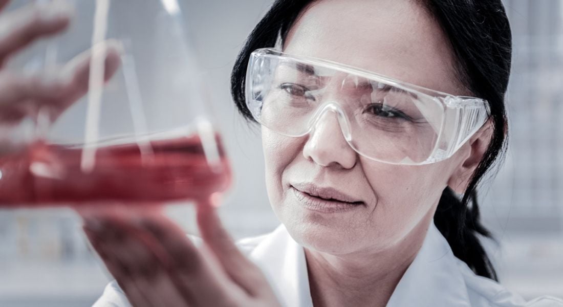 A close-up photo of a women holding up a scientific flask containing a red liquid in a lab setting.