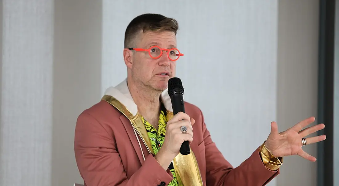 Man in highlighter orange framed glasses and salmon coloured blazer holding microphone and giving an address.