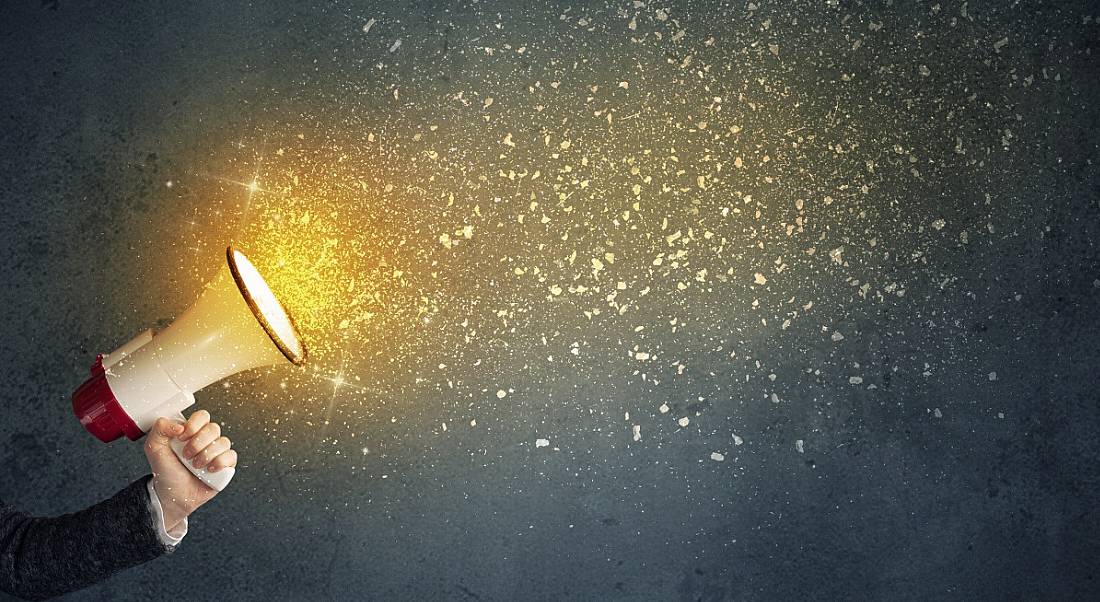 Hand holding a megaphone with glowing confetti coming out of it, against a dark background.