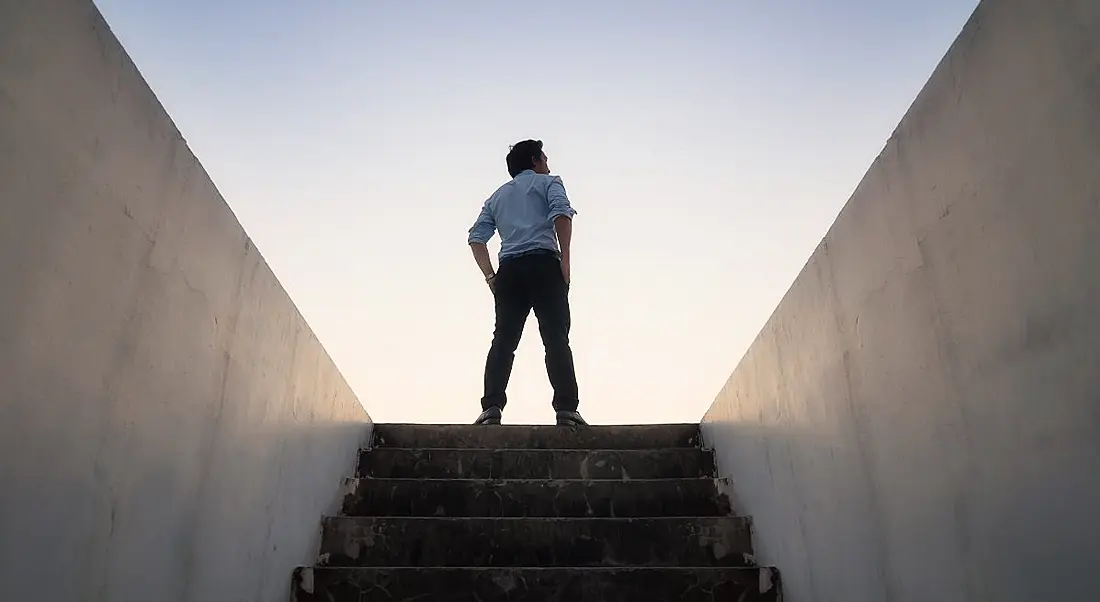 Person standing confidently at the top of a staircase to outside against a setting sun.