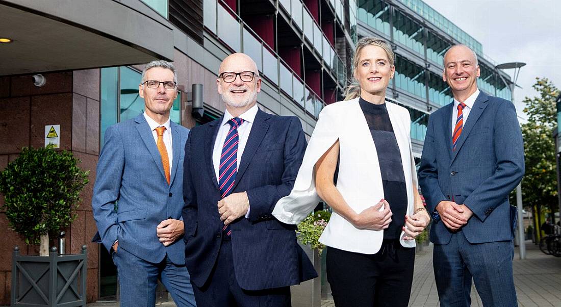 Three businessmen and a businesswoman are facing the camera, standing in front of a glass-panelled office building.