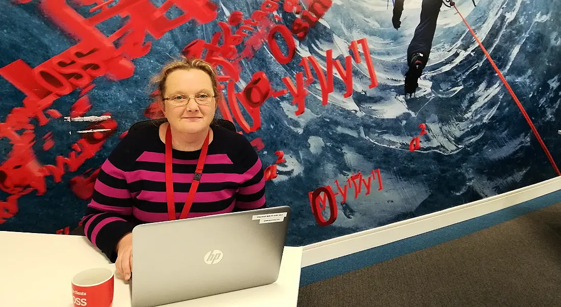 A blonde woman with glasses and a pink and black striped sweater sits in front of a laptop and looks at the camera.