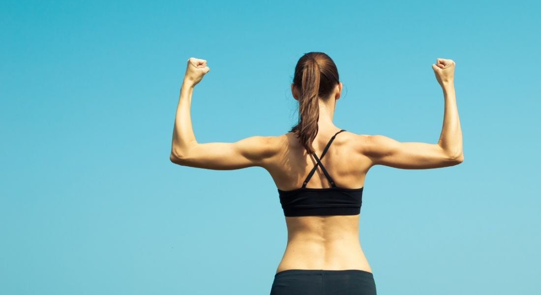 A fit woman in a sports bra and leggings flexing her muscles with her back to the camera, shot against a clear blue sky.