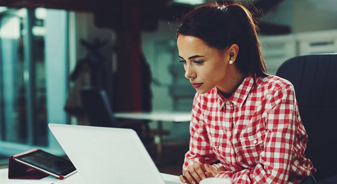 A woman in a red checked shirt sitting at a desk and staring intently at her laptop while working.