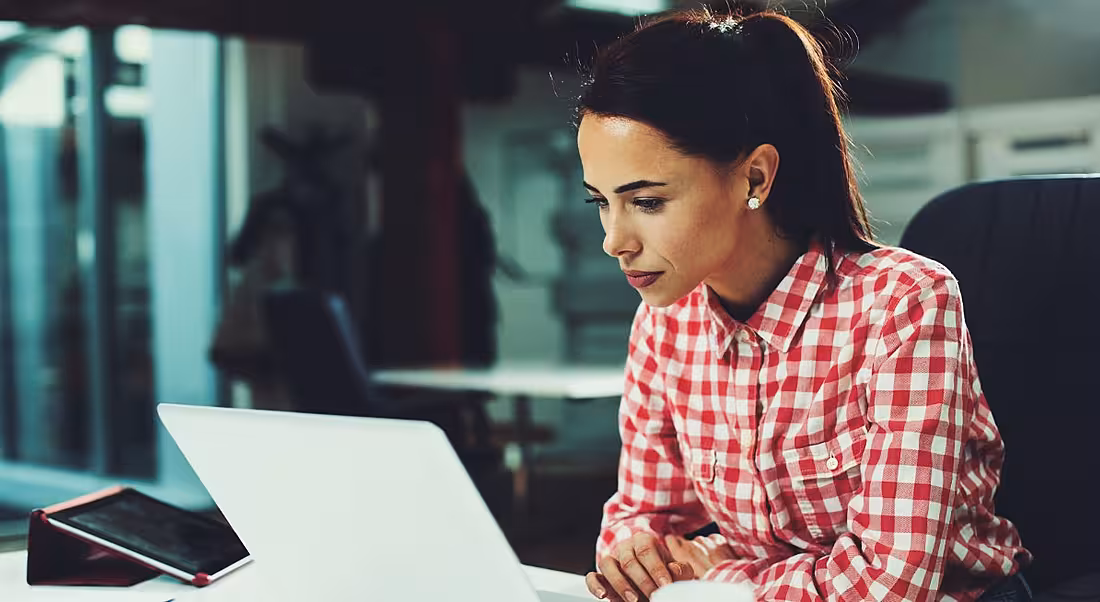 A woman in a red checked shirt sitting at a desk and staring intently at her laptop while working.