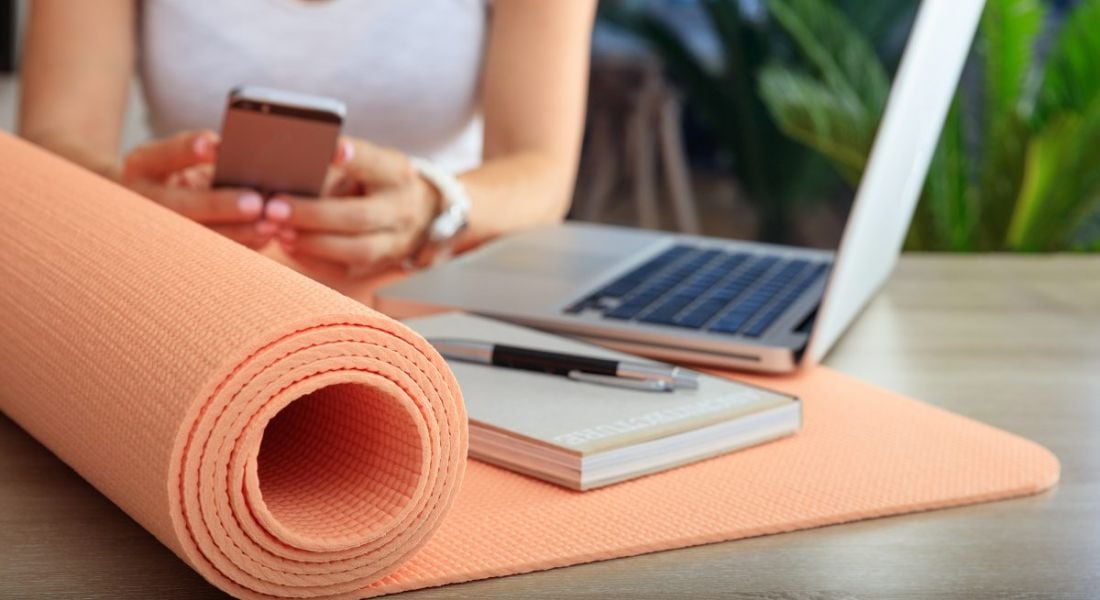 A woman in a white T-shirt texting on her phone with an orange yoga mat, a notebook and pen and an open laptop sitting on the table in front of her.