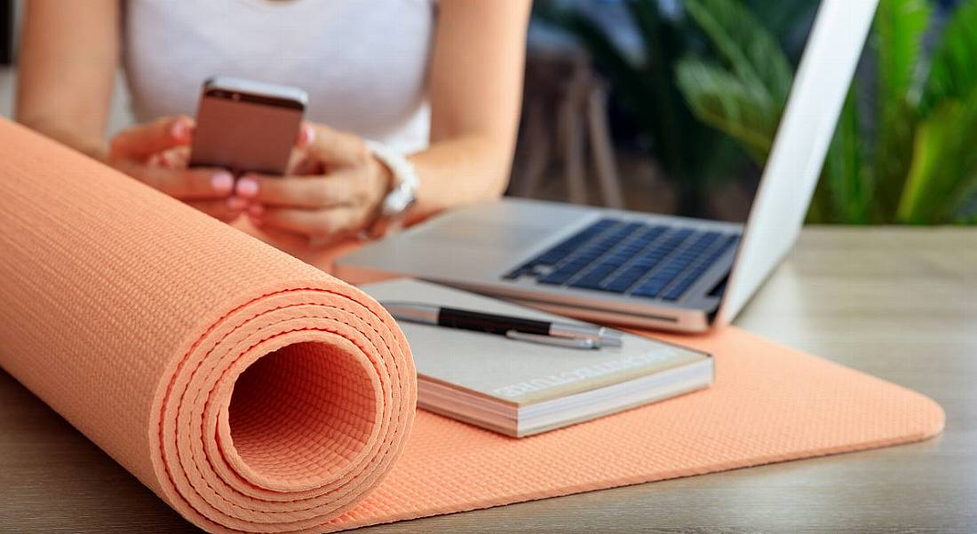 A woman in a white T-shirt texting on her phone with an orange yoga mat, a notebook and pen and an open laptop sitting on the table in front of her.