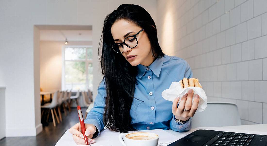 A woman working through lunch. She’s holding a sandwich in one hand and writing notes with the other in front of a laptop.