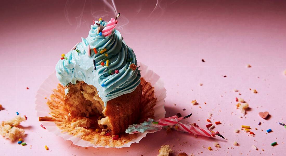 A gouged birthday cupcake with an extinguished candle sitting lopsided in the icing against a pink background.