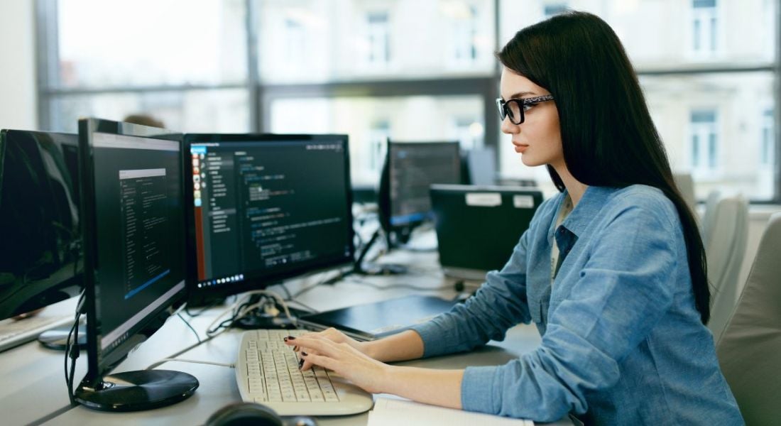 A woman at a computer with two screens showing code against a dark background. She’s using a programming language.