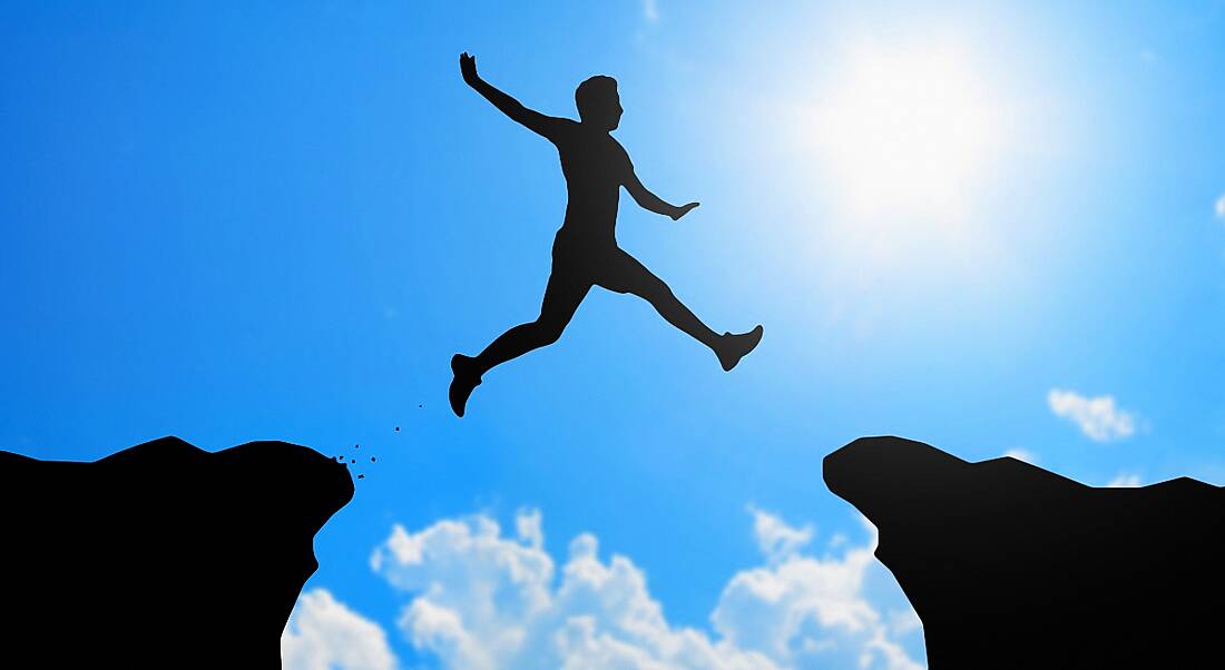 A silhouette of a man jumping from one cliff to another against blue sky to represent job-hopping.