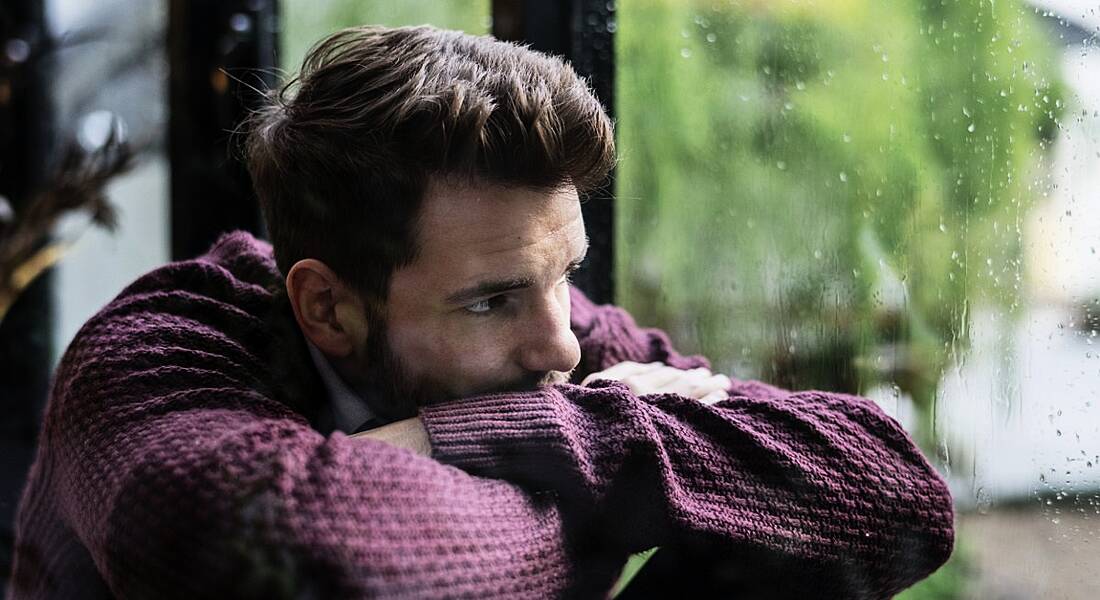 A man in a jumper with his head resting on his arms looking out the window at the rain. It’s Blue Monday and he looks sad.