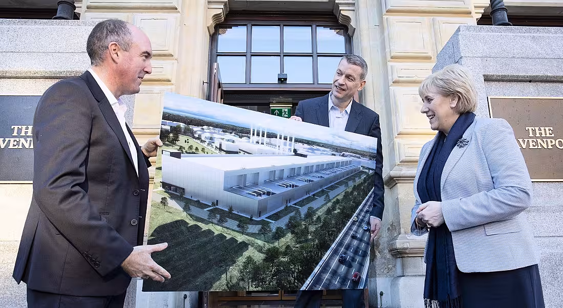 View of three middle aged people bundled in scarves and coats loosely holding up a large printout of a data centre.