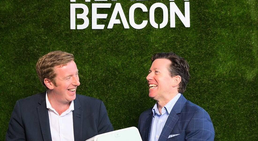 Two men in dark jackets sit before a green wall holding a white medical device in front of a green wall with HealthBeacon branding in white.