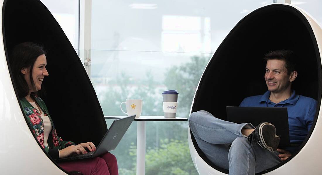Two Globoforce employees, one male and one female, sitting in egg chairs facing each other. They have laptops on their lap.