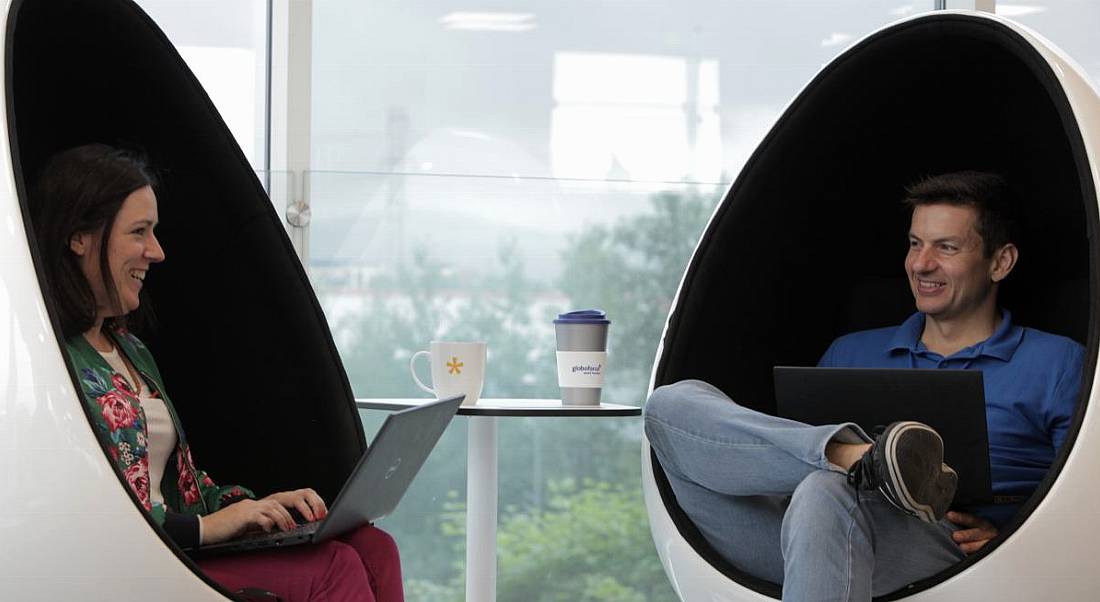 Two Globoforce employees, one male and one female, sitting in egg chairs facing each other. They have laptops on their lap.