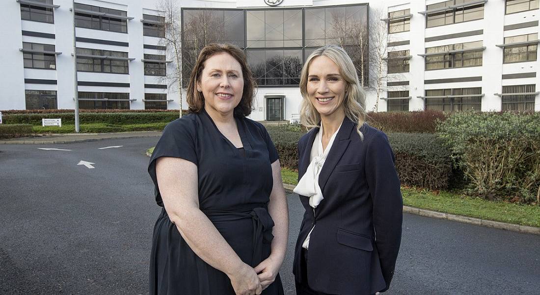 Two women in dark business attire smiling at camera against backdrop of Three Ireland building in Limerick.