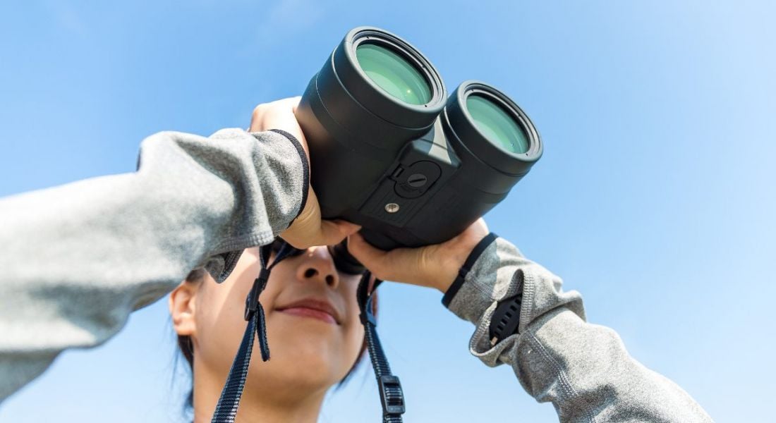 A close-up of a woman with binoculars against a blue-sky backdrop. It represents looking ahead to workplace trends in 2019.