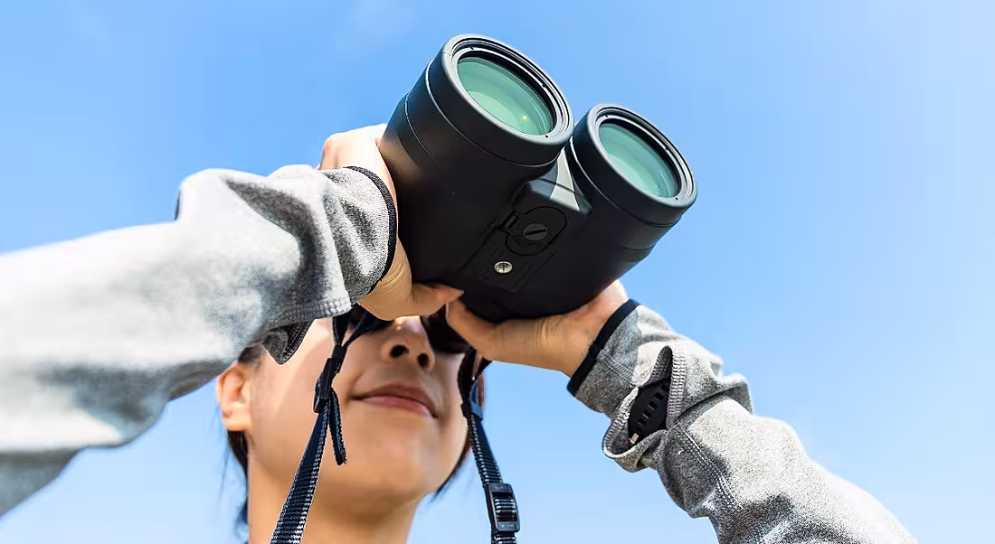 A close-up of a woman with binoculars against a blue-sky backdrop. It represents looking ahead to workplace trends in 2019.