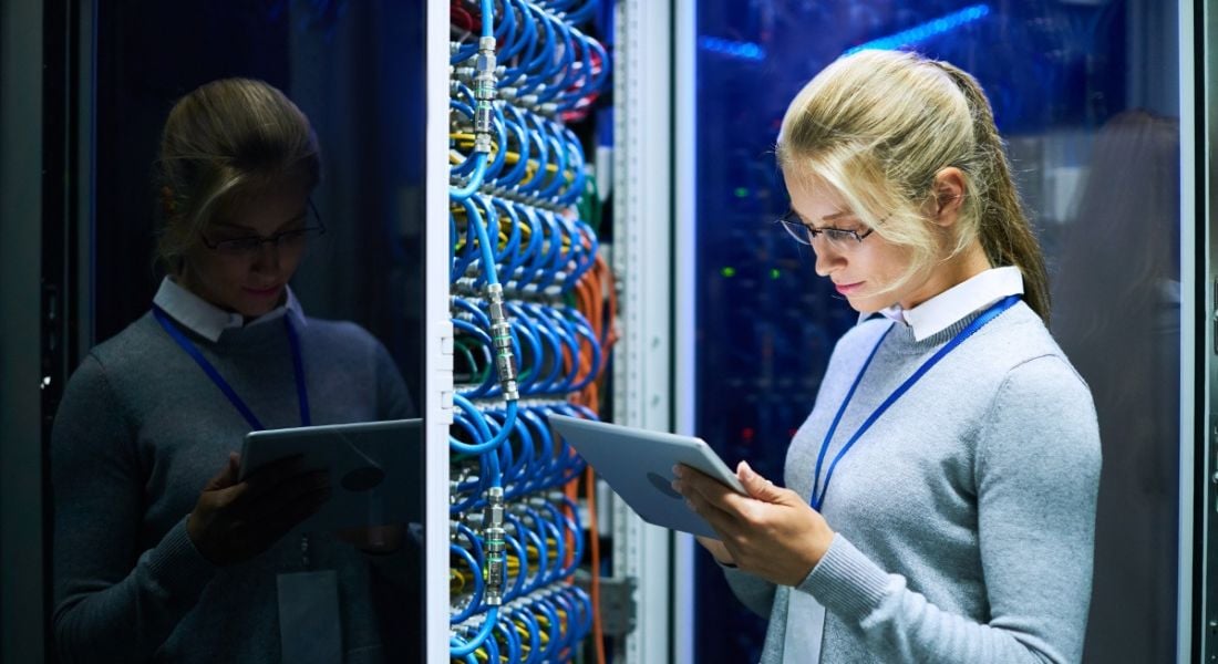 Blonde, female mainframe worker checking a tablet while standing beside a supercomputer full of blue wires.