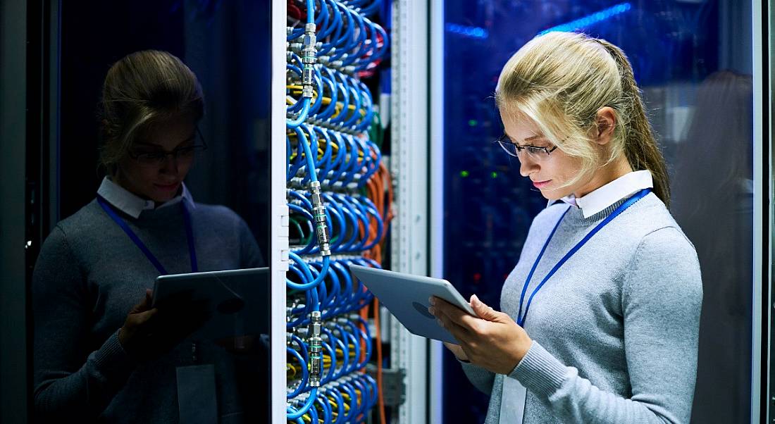 Blonde, female mainframe worker checking a tablet while standing beside a supercomputer full of blue wires.