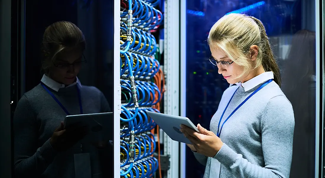 Blonde, female mainframe worker checking a tablet while standing beside a supercomputer full of blue wires.