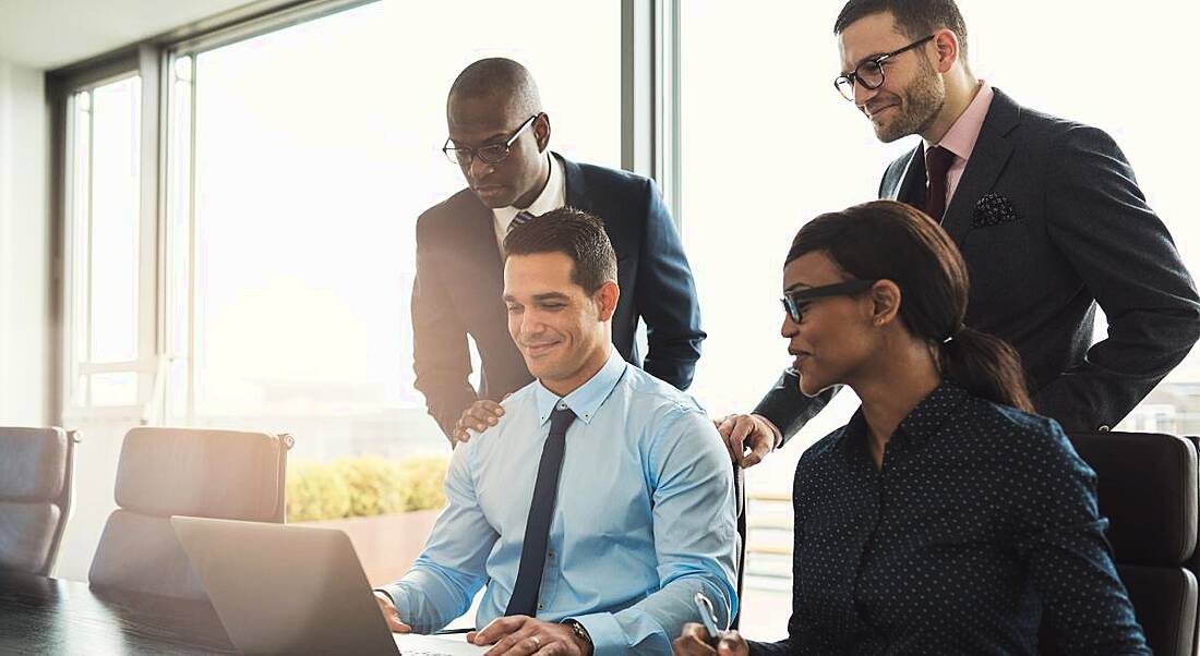 A diverse group of employees in a boardroom smiling and looking at a laptop.