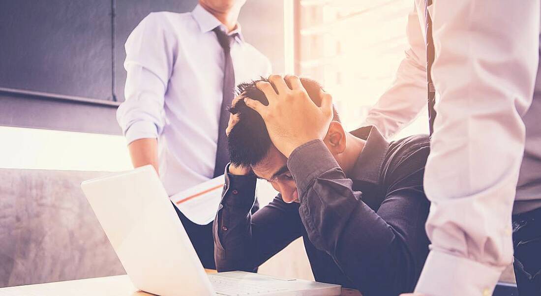 An employee at a laptop with his head in his hands while two colleagues stand over him, depicting workplace harassment.