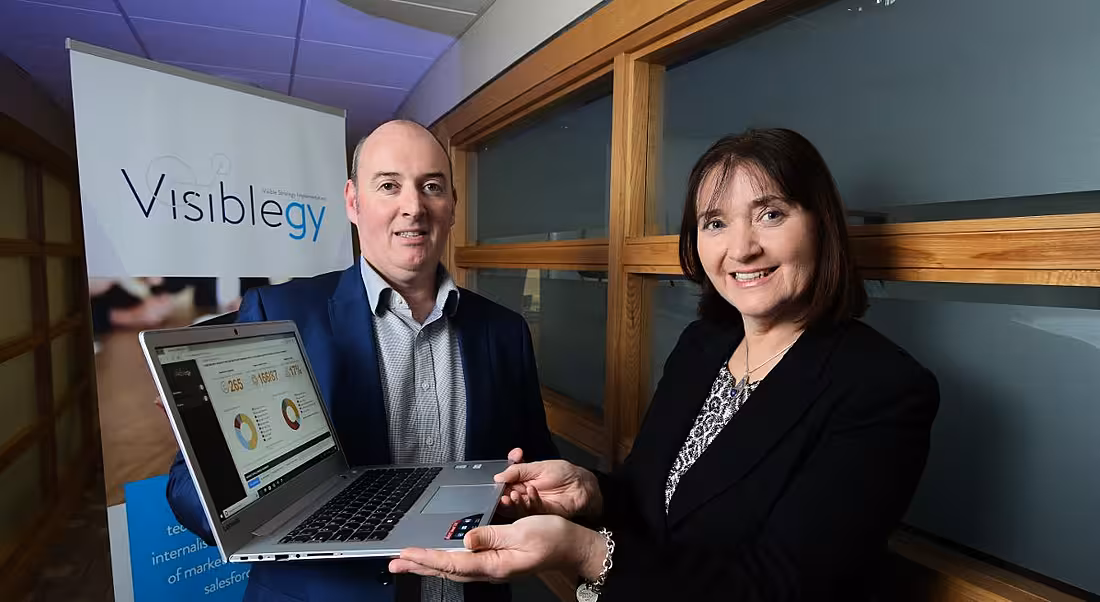 Image of a man and a woman posing awkwardly with a laptop in front of a Visiblegy logo.