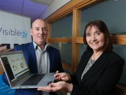 Formally dressed middle-aged men and woman standing in front of a purple wall with an Expleo logo.