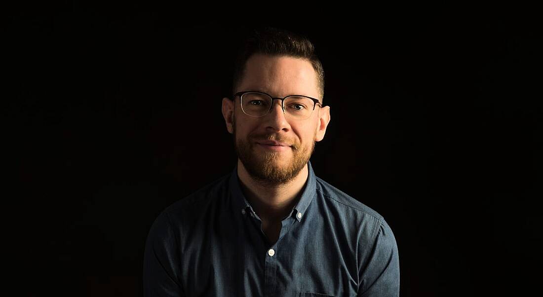 A young man in a blue shirt with horn-rimmed glasses smiles at the camera against a black background.