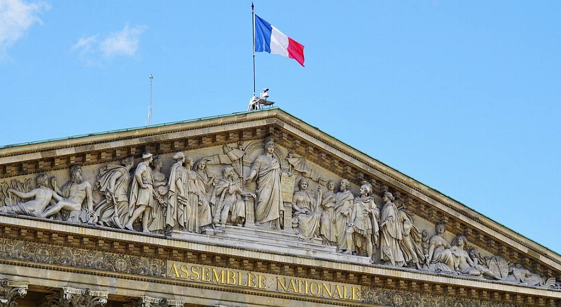 A view of an ornate government building In France with a French flag flying overhead against a clear sky.