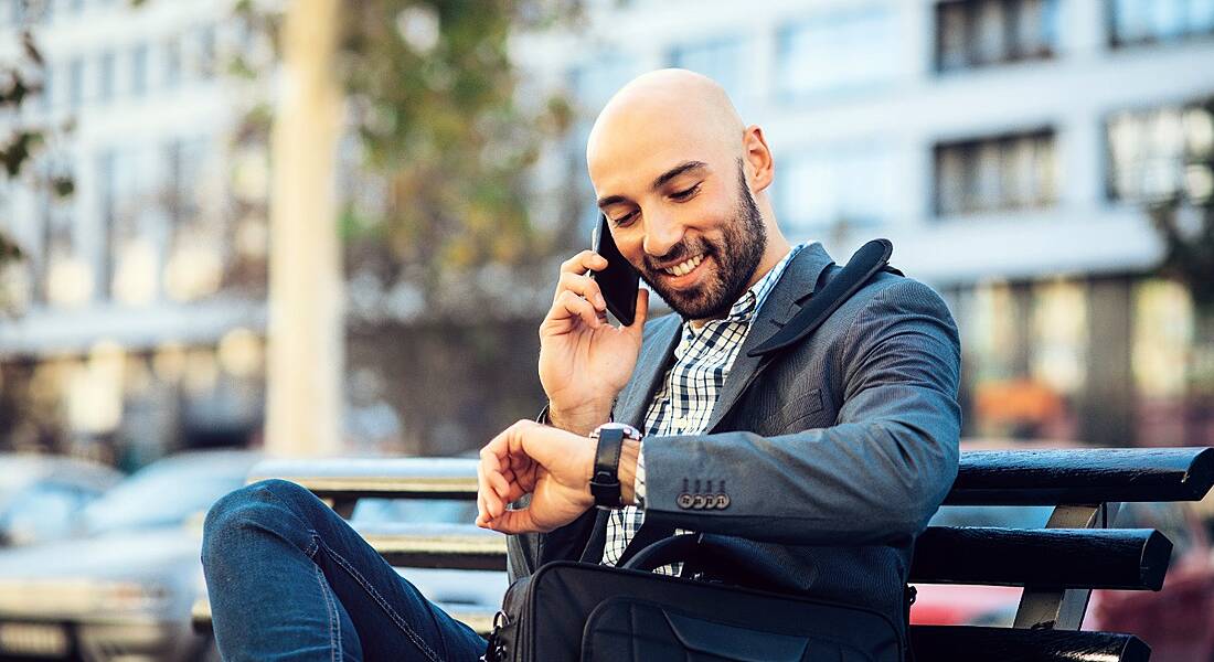 A young businessman sitting on a park bench n a metropolitan area looking at his watch and smiling.