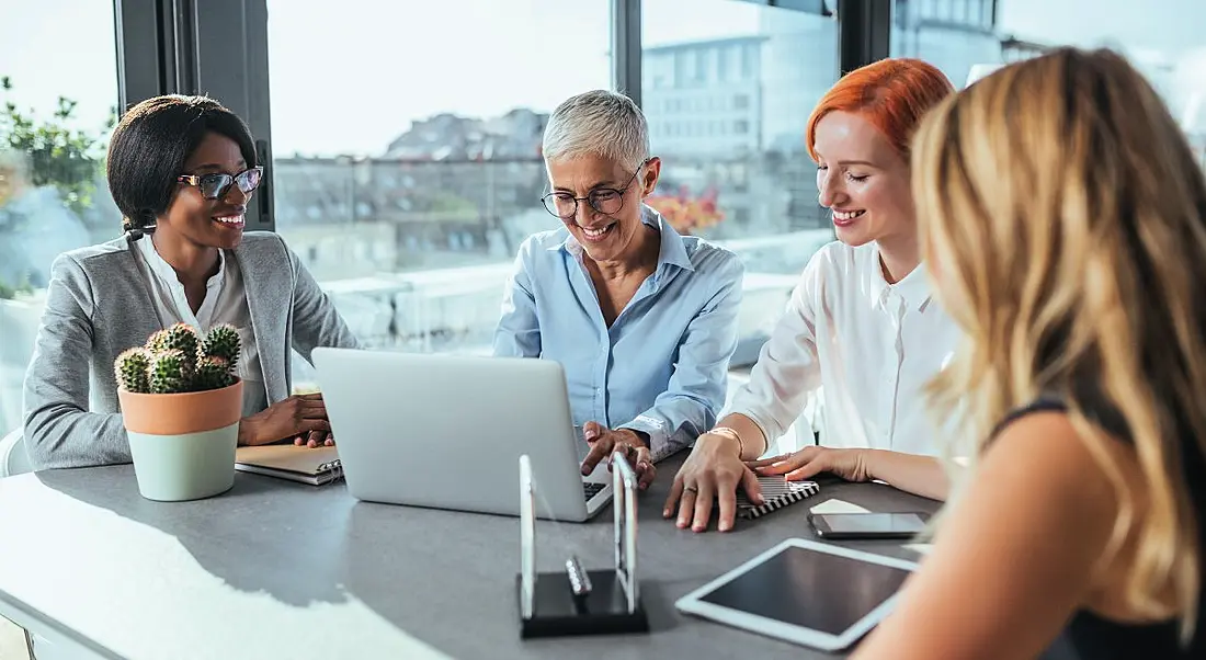 Four women of different ages work together on devices and notebooks in a meeting room overlooking a cityscape.