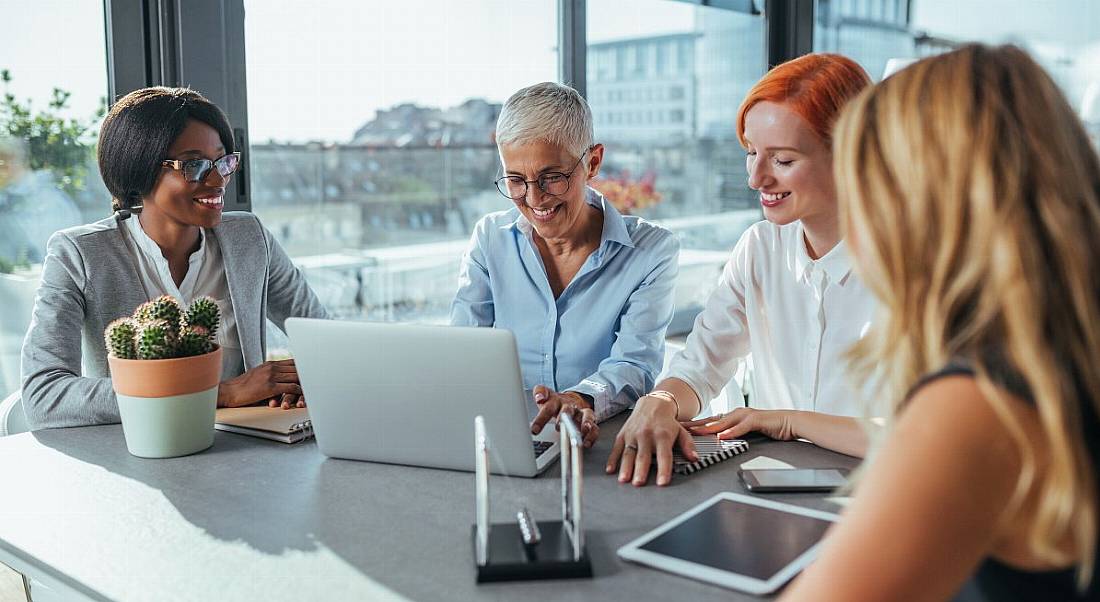 Four women of different ages work together on devices and notebooks in a meeting room overlooking a cityscape.