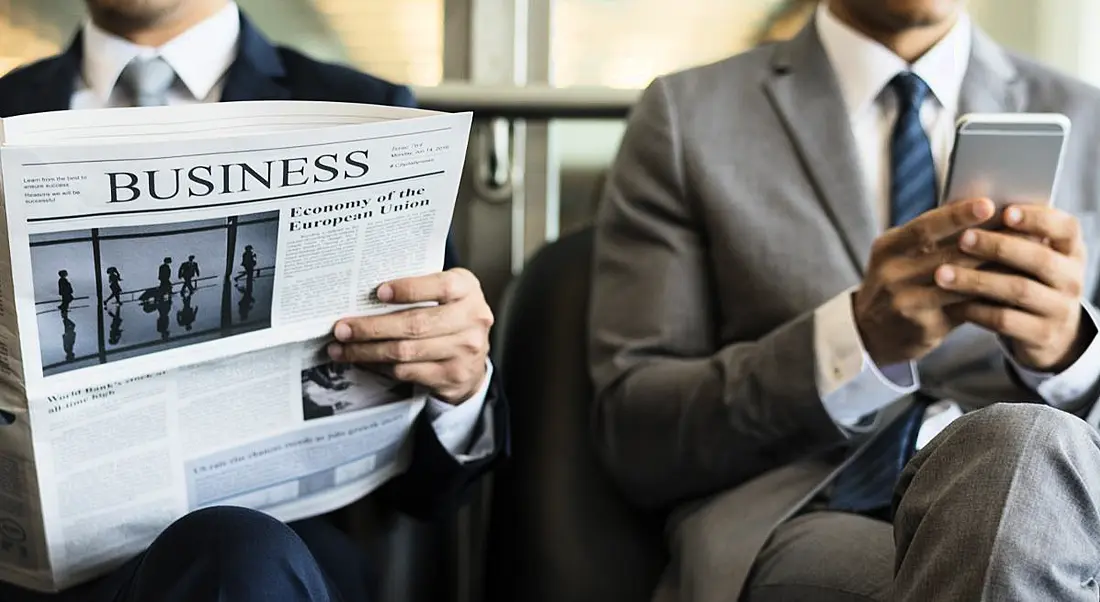 Two men in suits, the one on the left reading the business section and the one on the right using his phone.