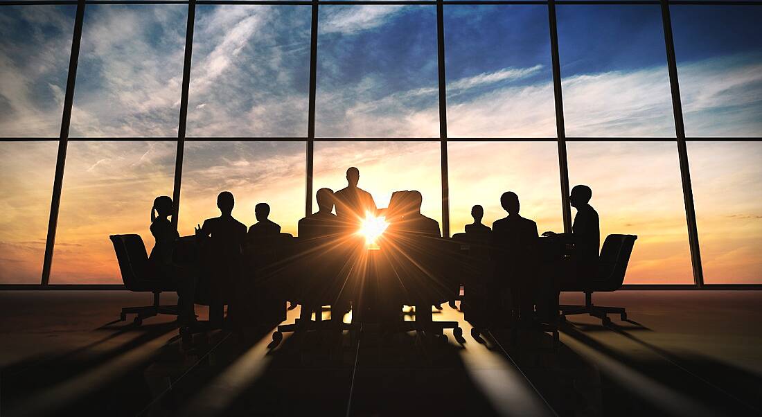 Silhouette of a team of people at a meeting in a room with wall-to-ceiling glass through which the sunrise is streaming in.