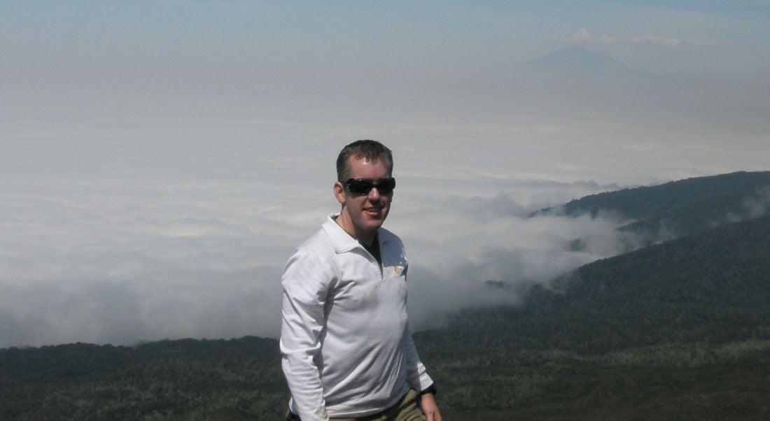 A smiling man wearing sunglasses, standing in front of a mountain landscape. He works for Johnson & Johnson Vision Care.